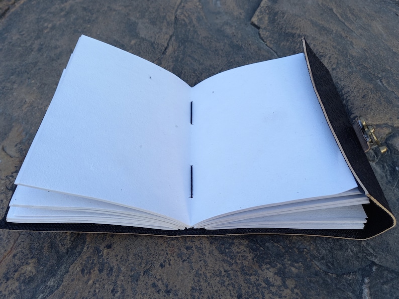 Op de afbeelding: Een zwart lederen gebonden dagboek met een messing sluiting, open om blanco witte pagina's te onthullen die met blauwe draad aan elkaar zijn genaaid.