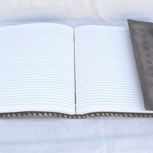 K&ouml;nnte beinhalten: Ein braunes, ledergebundenes Tagebuch mit einem dekorativen Lesezeichen. Das Tagebuch hat linierte Seiten und einen schwarz-wei&szlig;en Musterrand.