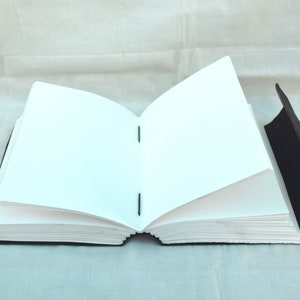 May include: An open, blank journal with a black leather cover and metal clasp. The pages are white and the binding is visible. The journal is lying open, displaying the blank pages, ready for writing or sketching.