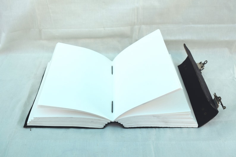 May include: An open, blank journal with a black leather cover and metal clasp. The pages are white and the binding is visible. The journal is lying open, displaying the blank pages, ready for writing or sketching.
