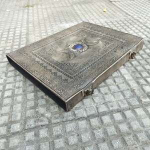 May include: A gray leather journal with an intricate design and a blue gemstone in the center. The journal has a clasp closure and is perfect for writing or keeping secrets.