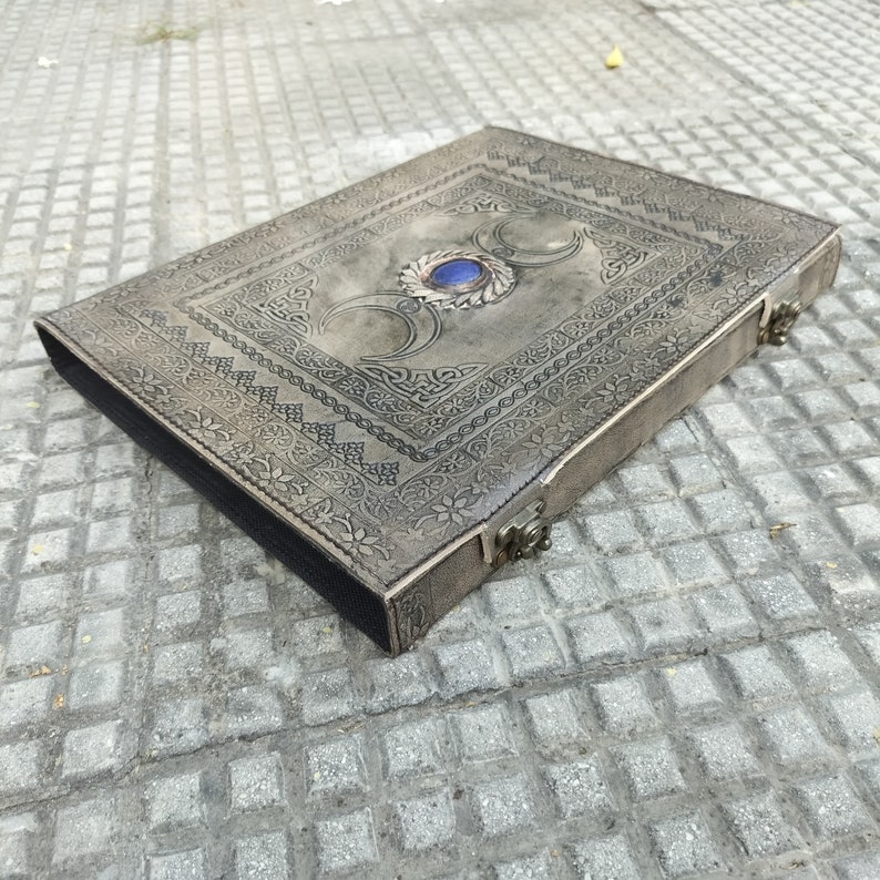 May include: A gray leather journal with an intricate design and a blue gemstone in the center. The journal has a clasp closure and is perfect for writing or keeping secrets.