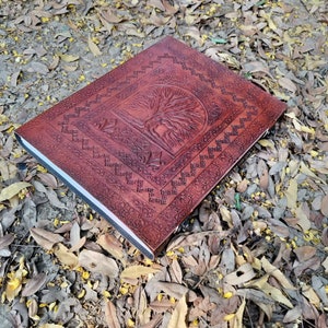 Puede incluir: Un diario de cuero marr&oacute;n con un dise&ntilde;o de &aacute;rbol de la vida en relieve. El diario tiene un borde decorativo y est&aacute; sobre un lecho de hojas ca&iacute;das.