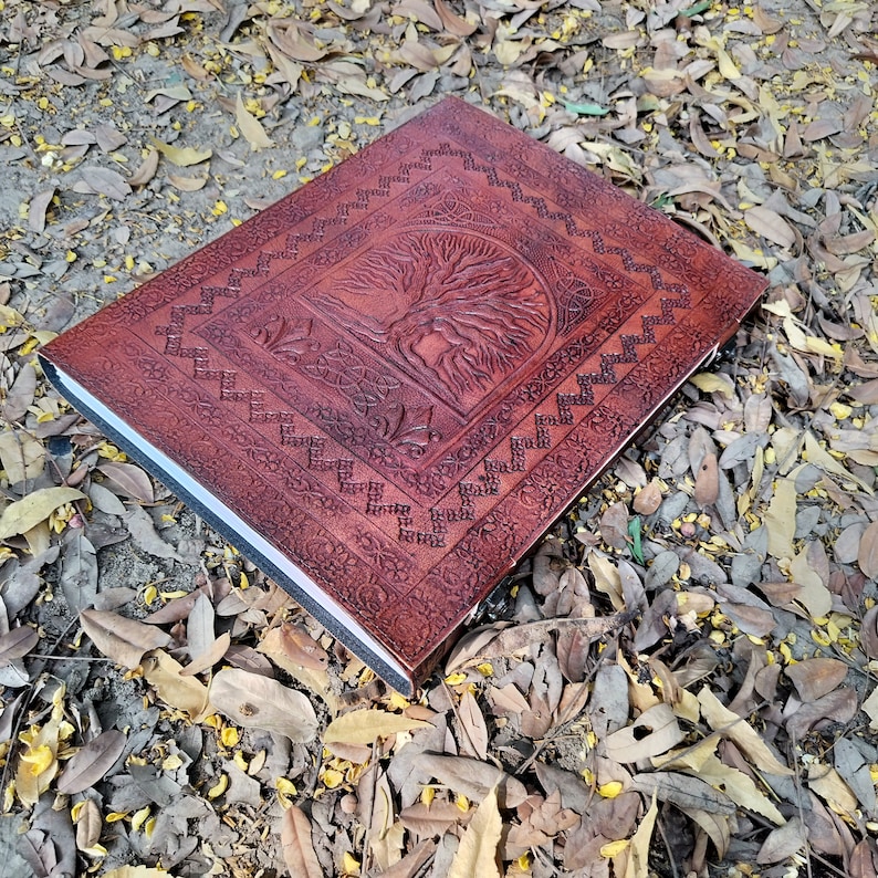 Puede incluir: Un diario de cuero marr&oacute;n con un dise&ntilde;o de &aacute;rbol de la vida en relieve. El diario tiene un borde decorativo y est&aacute; sobre un lecho de hojas ca&iacute;das.