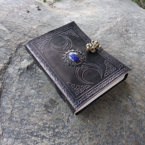 Op de afbeelding: Zwart lederen dagboek met een drievoudig maandesign en een blauwe edelsteen. Het dagboek heeft een metalen sluiting.
