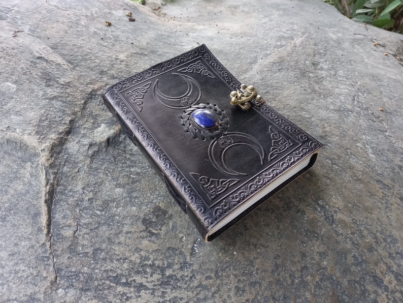 Op de afbeelding: Zwart lederen dagboek met een drievoudig maandesign en een blauwe edelsteen. Het dagboek heeft een metalen sluiting.