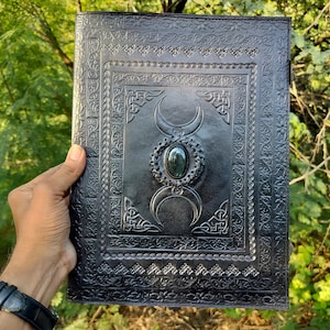 May include: Black leather journal with an embossed triple moon design and a green gemstone in the center. The journal has a decorative border around the edges.