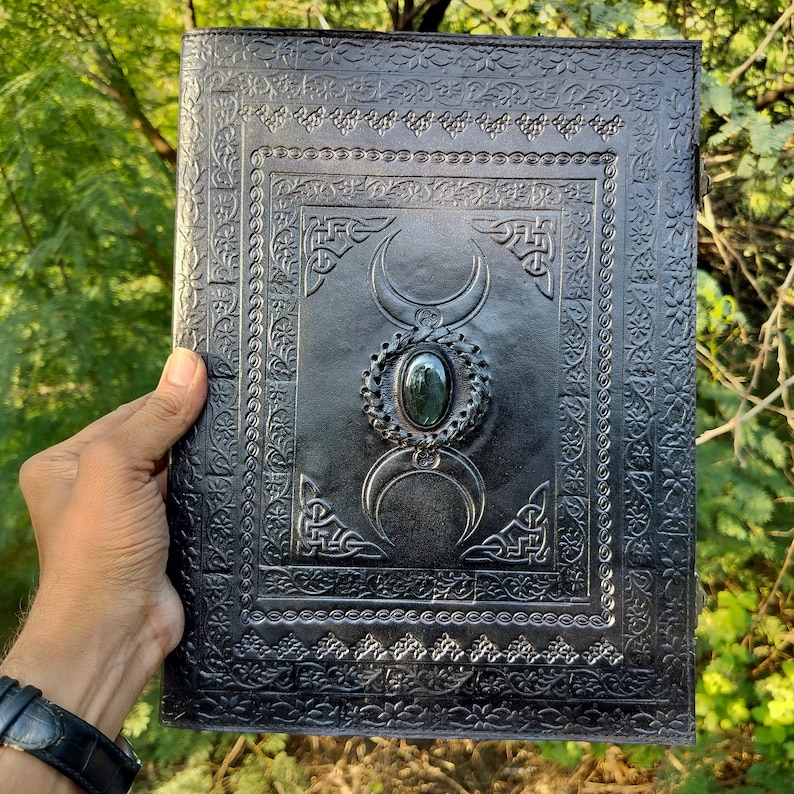 May include: Black leather journal with an embossed triple moon design and a green gemstone in the center. The journal has a decorative border around the edges.
