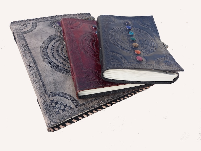 K&ouml;nnte beinhalten: Drei ledergebundene Journale mit aufwendigen Designs. Das obere Journal hat einen schwarzen Einband mit bunten Edelsteinen und einem Mandala-Muster. Das mittlere Journal hat einen burgunderfarbenen Einband mit einem &auml;hnlichen Mandala-Muster. Das untere Journal hat einen grauen Einband mit einem aufwendigeren Design.