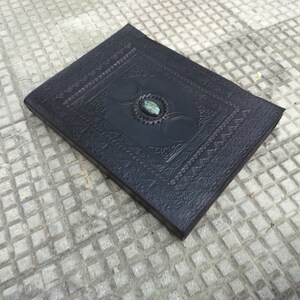 May include: A black leather journal with an intricate embossed design and a green gemstone in the center. The journal has a triple moon symbol on the front cover.