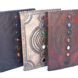 K&ouml;nnte beinhalten: Drei ledergebundene Journale mit dekorativen, gepr&auml;gten Designs und bunten Edelstein-Akzenten. Die Journale sind geschlossen und in einer Reihe angeordnet.