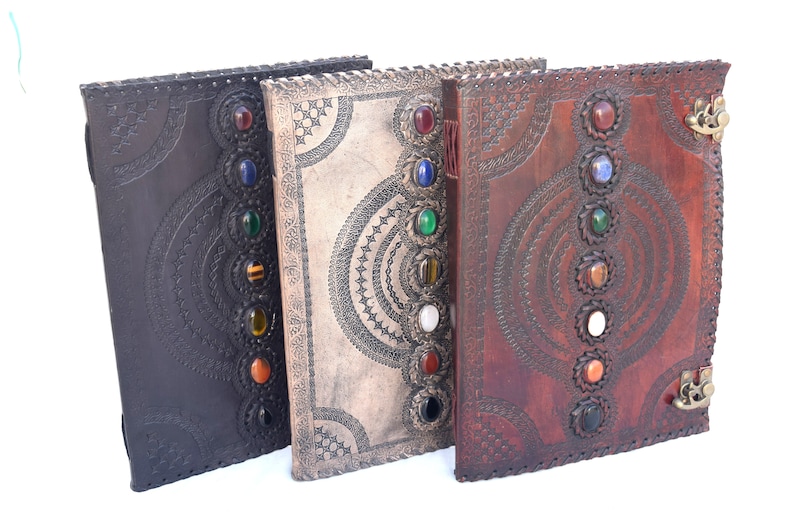 K&ouml;nnte beinhalten: Drei ledergebundene Journale mit dekorativen, gepr&auml;gten Designs und bunten Edelstein-Akzenten. Die Journale sind geschlossen und in einer Reihe angeordnet.