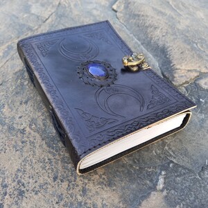 Op de afbeelding: Een donkerbruin leren dagboek met een decoratief reli&euml;fdesign met drie halve manen en een blauwe edelsteen. Het dagboek heeft een messing sluiting.