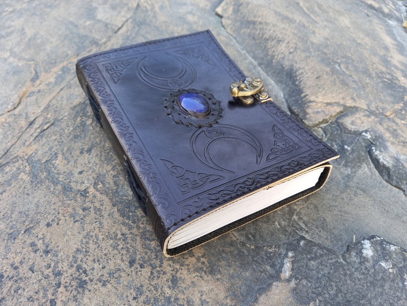 Op de afbeelding: Een donkerbruin leren dagboek met een decoratief reli&euml;fdesign met drie halve manen en een blauwe edelsteen. Het dagboek heeft een messing sluiting.