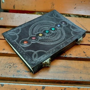 May include: A dark brown leather journal with a decorative embossed design and seven colorful gemstones set in the center. The journal has a clasp closure.