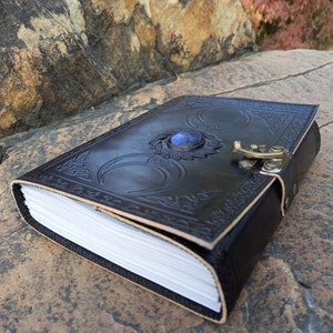 Op de afbeelding: Een zwart lederen dagboek met een drievoudig maandesign en een blauwe edelsteenaccent. Het dagboek heeft een sluiting met een gesp en is versierd met ingewikkelde details.