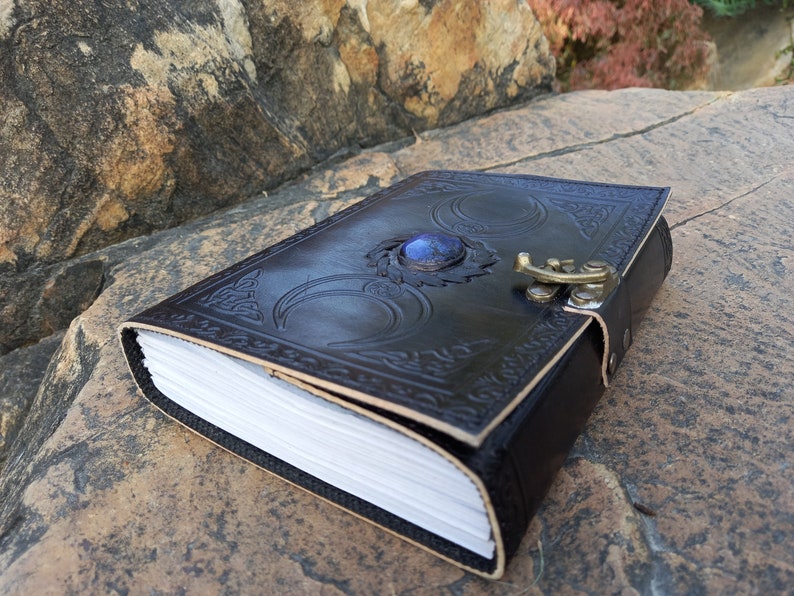 Op de afbeelding: Een zwart lederen dagboek met een drievoudig maandesign en een blauwe edelsteenaccent. Het dagboek heeft een sluiting met een gesp en is versierd met ingewikkelde details.