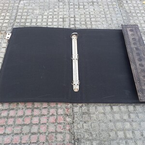 May include: A black binder with a silver ring mechanism and a decorative brown leather spine. The binder is open and lying flat on a gray tiled surface.