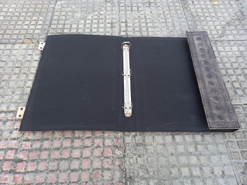 May include: A black binder with a silver ring mechanism and a decorative brown leather spine. The binder is open and lying flat on a gray tiled surface.