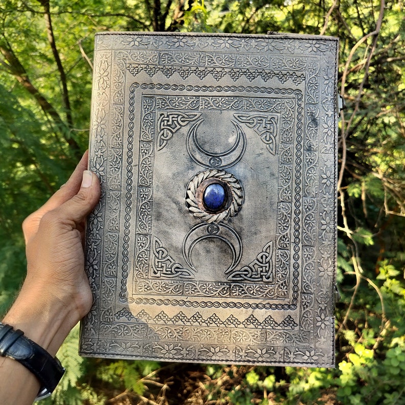May include: A silver leather journal with a decorative design featuring a triple moon symbol and a blue gemstone. The journal has a distressed look and is perfect for writing or journaling.