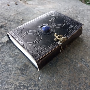 Op de afbeelding: Een zwart lederen dagboek met een decoratief reli&euml;fdesign van drie halve manen en een blauwe edelsteen. Het dagboek heeft een messing sluiting en een band.