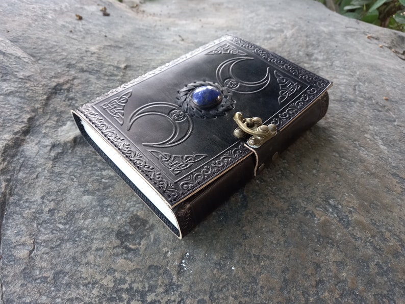 Op de afbeelding: Een zwart lederen dagboek met een decoratief reli&euml;fdesign van drie halve manen en een blauwe edelsteen. Het dagboek heeft een messing sluiting en een band.
