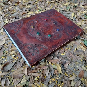 Pu&ograve; includere: Un diario in pelle marrone con un design a pentagramma e pietre preziose colorate. Il diario ha un bordo decorativo e una chiusura.