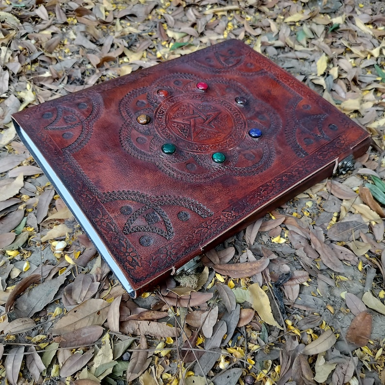 Pu&ograve; includere: Un diario in pelle marrone con un design a pentagramma e pietre preziose colorate. Il diario ha un bordo decorativo e una chiusura.