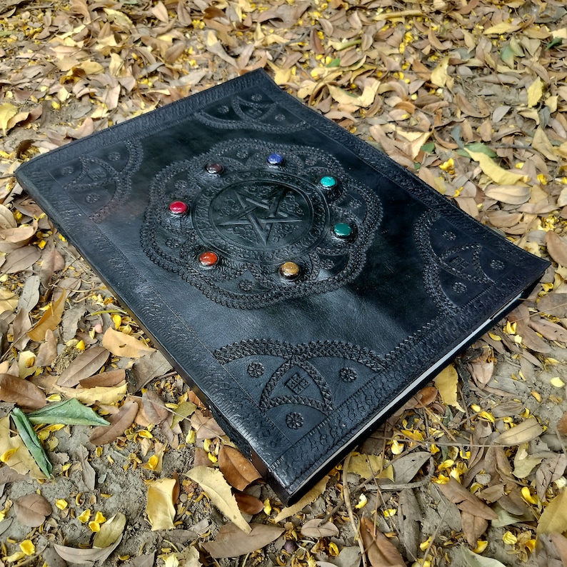 Pu&ograve; includere: Diario in pelle nera con un design a pentagramma e pietre preziose colorate attorno ai bordi. Il diario &egrave; sdraiato su un letto di foglie cadute.