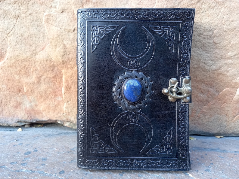 Op de afbeelding: Zwart lederen dagboek met een decoratief ontwerp met een halve maan en een lapis lazuli-steen. Het dagboek heeft een metalen sluiting.