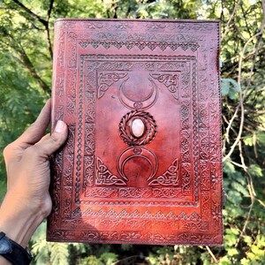 May include: A brown leather journal with an intricate embossed design featuring a triple moon symbol and a white stone. The journal has a Celtic knot border and is perfect for writing, journaling, or spell casting.
