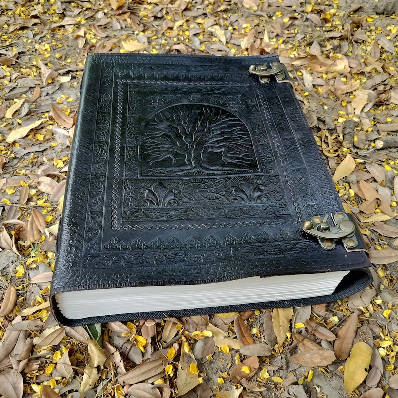 Puede incluir: Un diario de cuero negro con un dise&ntilde;o de &aacute;rbol de la vida en relieve en la portada. El diario tiene dos cierres met&aacute;licos y est&aacute; sobre un lecho de hojas marrones.