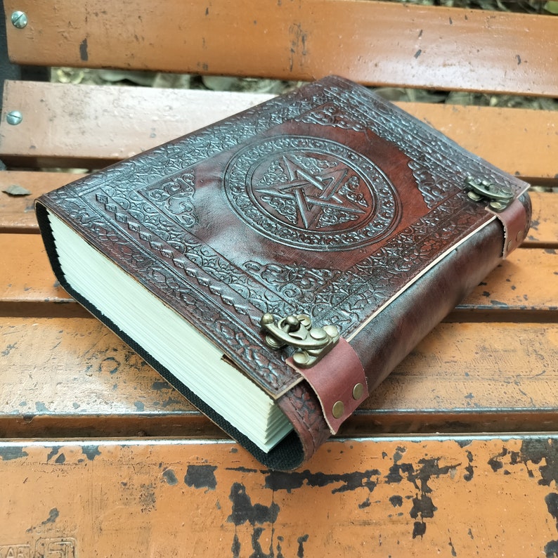 May include: A brown leather journal with a raised pentagram design on the cover. The journal has a clasp closure and a strap. The pages are cream-colored and the journal is sitting on a wooden surface.