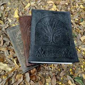 Puede incluir: Tres diarios encuadernados en cuero con dise&ntilde;os en relieve de &aacute;rbol de la vida. Los diarios est&aacute;n apilados uno encima del otro, con el diario negro en la parte superior, un diario marr&oacute;n en el medio y un diario gris en la parte inferior.