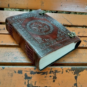 May include: A brown leather journal with a pentagram design embossed on the cover. The journal has a decorative border, metal clasps, and a burgundy spine. The pages are white and the journal is closed.