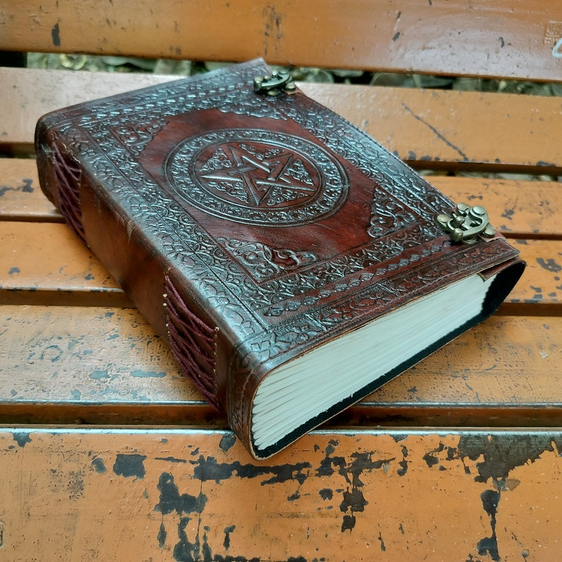 May include: A brown leather journal with a pentagram design embossed on the cover. The journal has a decorative border, metal clasps, and a burgundy spine. The pages are white and the journal is closed.