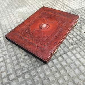 May include: A brown leather journal with a triple moon design and a pink stone in the center. The journal has a clasp closure and intricate embossed details.