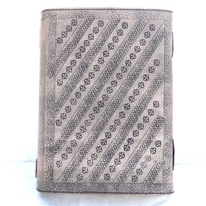 May include: A rectangular, light gray journal with a detailed, embossed pattern. The design features diagonal rows of floral and diamond shapes. The edges have a decorative border. The journal is standing upright against a white background.