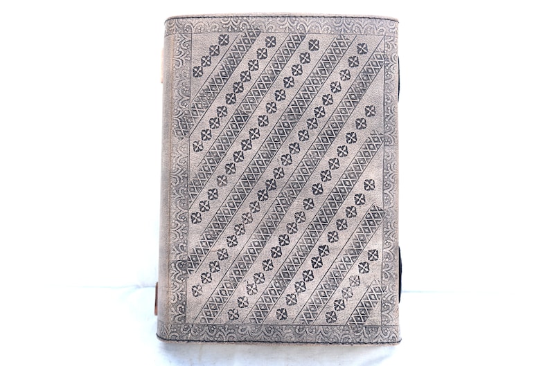 May include: A rectangular, light gray journal with a detailed, embossed pattern. The design features diagonal rows of floral and diamond shapes. The edges have a decorative border. The journal is standing upright against a white background.