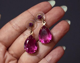 May include: A pair of gold-toned drop earrings with large, teardrop-shaped, pink gemstones. Each earring has a smaller, round pink gem at the top. The earrings are displayed against a dark gray background.