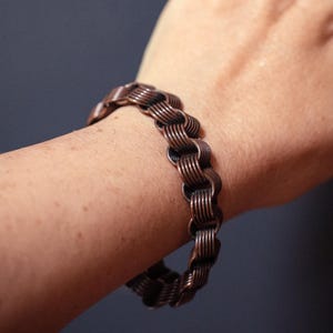 Puede incluir: Una pulsera de color cobre con un diseño de cadena trenzada. La pulsera está hecha de múltiples eslabones pequeños e interconectados, creando una superficie texturizada. La joya se lleva en una muñeca sobre un fondo oscuro.