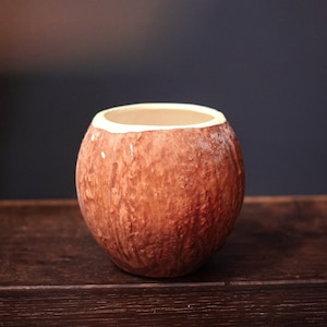 May include: A brown coconut shell cup with a white rim. The cup is sitting on a dark brown wooden surface.