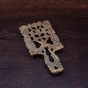 Könnte beinhalten: Ein goldfarbener Metall-Topfuntersetzer mit einem filigranen Design, das einen Baum und florale Motive zeigt. Der Topfuntersetzer liegt auf einer dunkelbraunen Holzoberfläche.