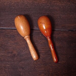 May include: Two wooden maracas with smooth, rounded heads and slender handles. The left maraca features a light brown, natural wood grain finish, while the right maraca has a slightly darker, polished reddish-brown tone with visible wood texture.