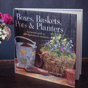 Puede incluir: Un libro titulado "Boxes, Baskets, Pots & Planters: A practical guide to 100 inspirational containers" de Stephanie Donaldson. La portada del libro presenta un fondo de madera rústico con un cubo de metal, una cesta tejida llena de flores y una pequeña maceta de terracota.
