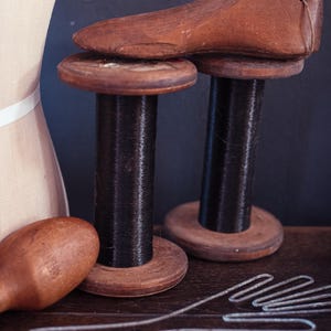 May include: A wooden shoe last resting on a dark brown wooden spool, accompanied by a second empty spool, a small wooden darning egg, a wire hand-shaped form, and a partial view of a beige fabric dress form with a white horizontal band.