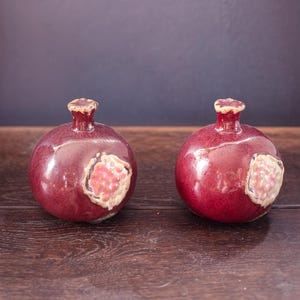 Peut inclure: Deux ornements en céramique en forme de grenade avec une finition rouge profond brillante. Chaque pièce présente une petite tige sur le dessus et une ouverture texturée de couleur claire sur le côté révélant des détails en forme de graines roses.