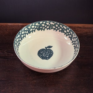 Puede incluir: Un cuenco de cerámica blanco con un diseño de manzana azul oscuro en el centro. El borde presenta un patrón de puntos en el mismo color azul oscuro. El cuenco está sobre una superficie de madera oscura contra un fondo oscuro.