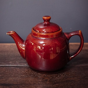 May include: A glossy, reddish-brown ceramic teapot with a lid and handle. The teapot is set on a wooden surface against a dark gray background. The teapot's design is simple and classic.