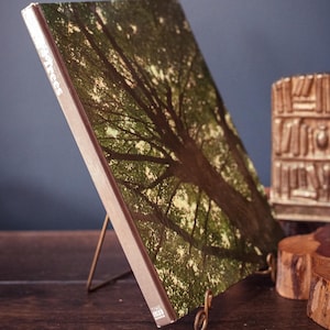 May include: A book titled "Trees" with a photograph of a tree canopy, displayed on a brass stand. The book's cover features a detailed image of tree branches and leaves. The book is propped up on a wooden surface.